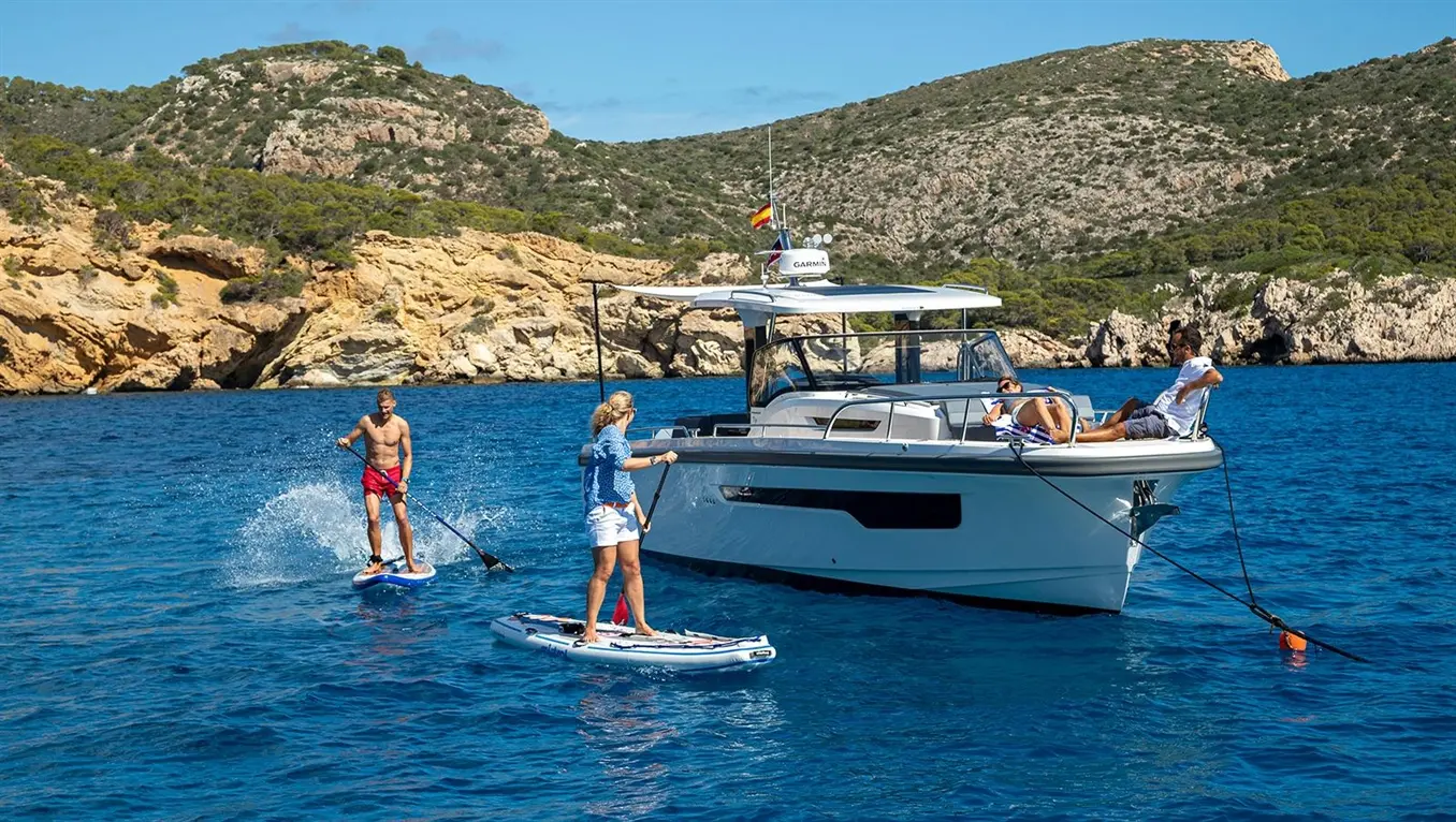 Two people paddleboarding near a boat in clear blue water, with rocky cliffs in the background, enjoying a sunny day.